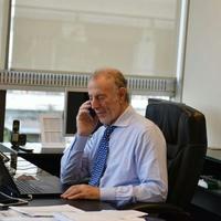 Un señor de edad avanzada vestido con ropa formal, camisa y corbata hablando por teléfono en una oficina